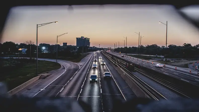 An interstate at dusk seen through a windshield, street lamps lit over the lanes